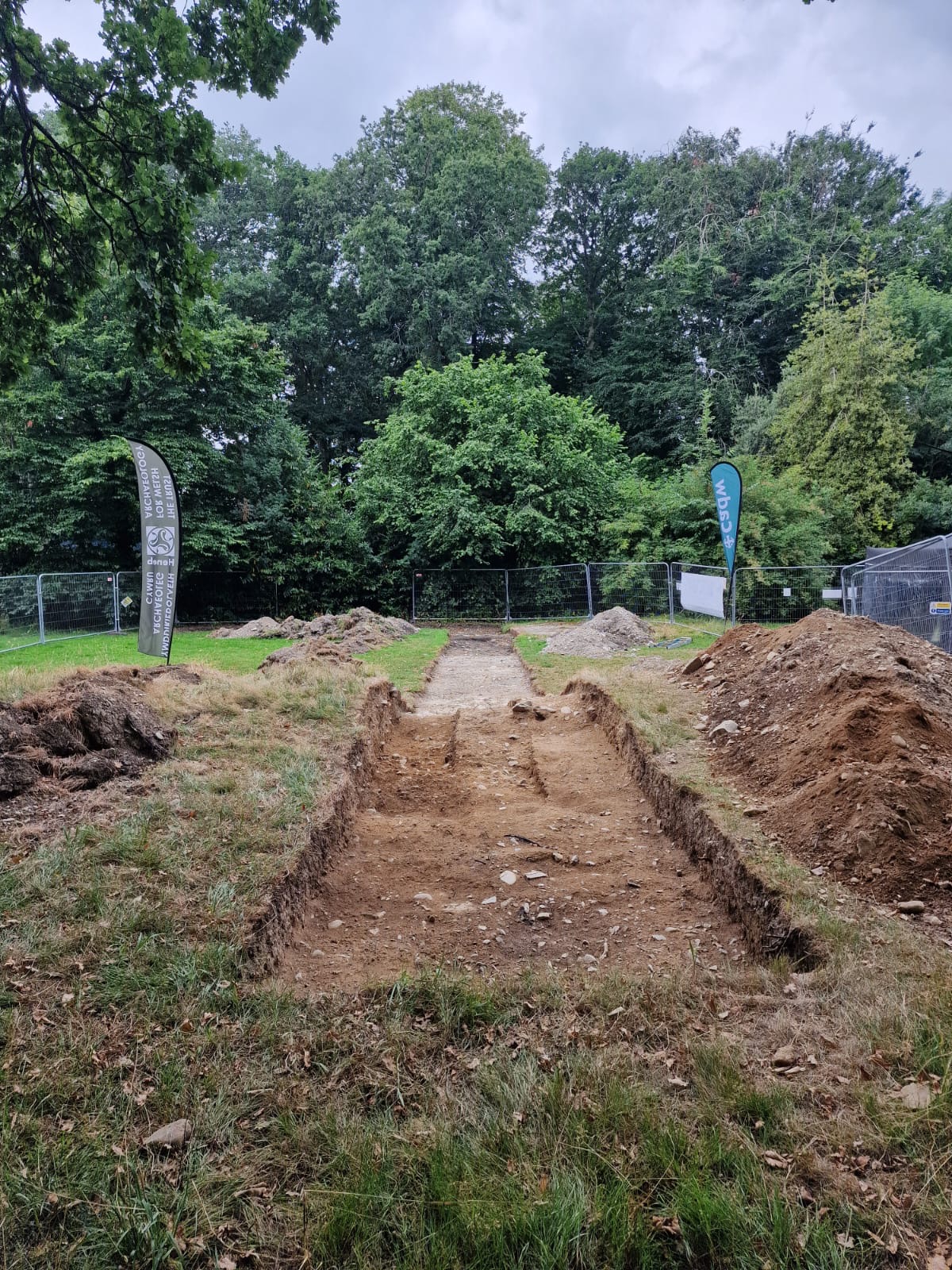 View of an open archaeological trench in a grassy area, with spoil heap on one side and cut turf on the other side and trees in the background, marked by informational flags and fencing.