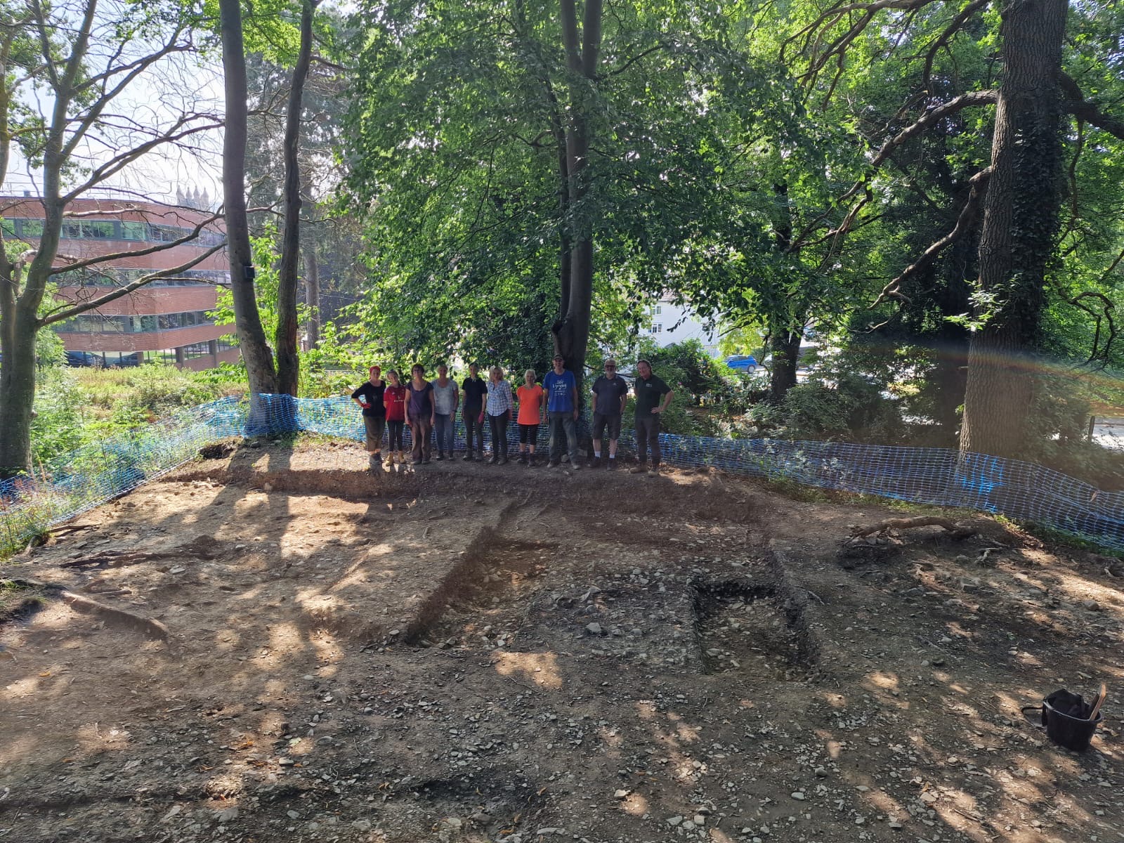 Group of archaeologists standing in front of an excavation area, surrounded by trees and blue safety fencing on a woodland site.