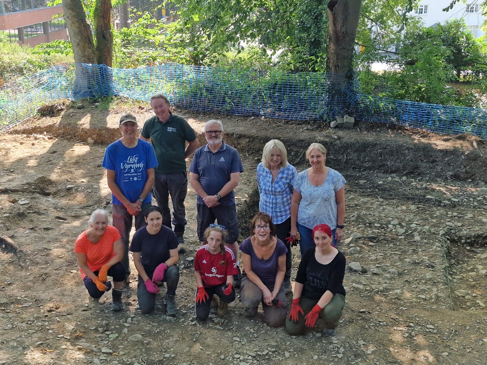 Smiling group of archaeology volunteers and staff posing at an excavation site in woodland, with visible trench edges and blue mesh safety fencing in the background