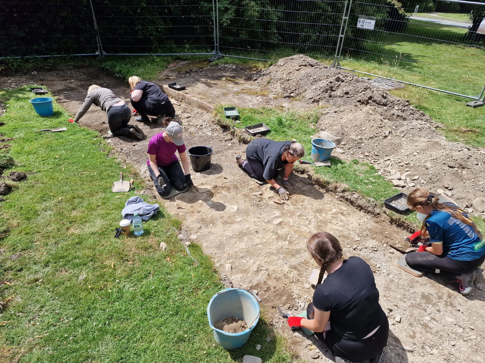 Group of archaeologists excavating a trench, using trowels and buckets, with fencing and trees in the background.
