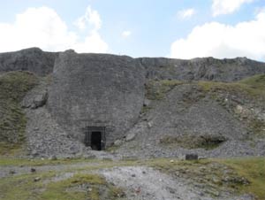 A large stone lime kiln built into a hillside, with a dark rectangular entrance at the base and loose rubble sloping down on either side under a partly cloudy sky