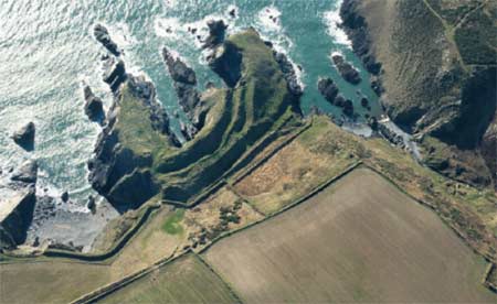 Oblique aerial view of a coastal promontory with steep rocky cliffs, earthwork defences, and enclosed fields overlooking the sea