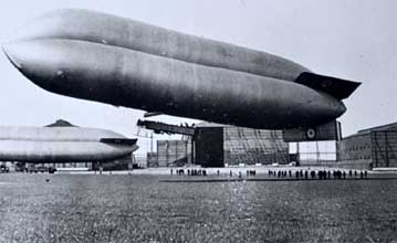 Historic black-and-white photograph showing a large airship lifting off near a hangar, with another airship and groups of people visible on the ground.