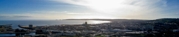 Panoramic view of a coastal city with the bay, harbour and surrounding hills visible under bright sunlight.