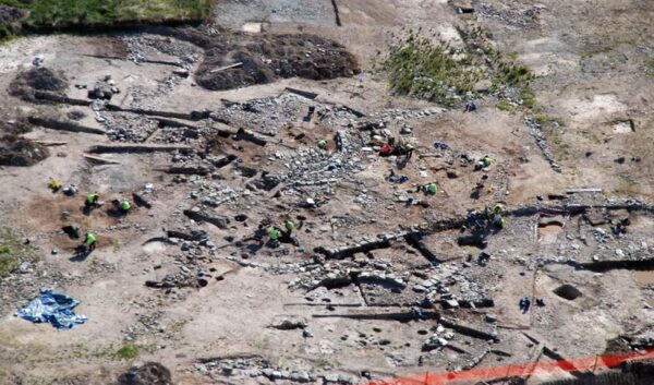 Aerial view of an archaeological excavation showing multiple roundhouse foundations and stone-built features, with archaeologists working across the site.