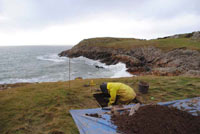 Archaeologist excavating a small test pit on a grassy coastal cliff edge with the sea and rocky shoreline in the background.