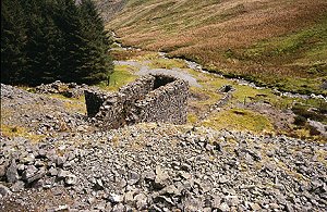 Ruined stone buildings and spoil heaps in a remote upland valley, with scattered rocks in the foreground,