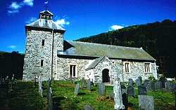 Stone-built rural church with a square tower and slate roof, surrounded by old gravestones in a grassy churchyard, set against a backdrop of wooded hills under a bright blue sky.