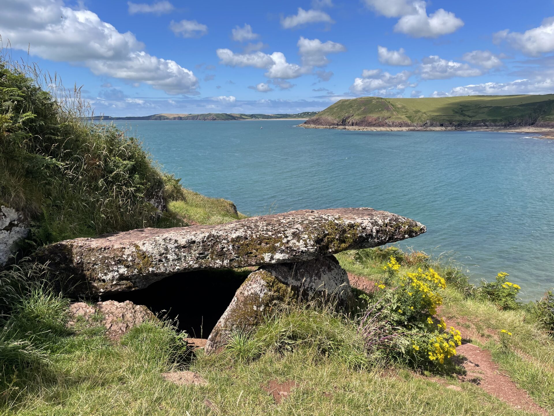 An ancient stone structure stands on a grassy hillside overlooking a serene coastline. The blue sea stretches into the distance, with hills and cliffs visible in the background under a partly cloudy sky. Yellow wildflowers grow near the dolmen, adding a splash of colour to the scene.