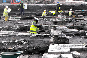 Archaeologists wearing high-visibility vests and hard hats excavating exposed structural remains on an active dig site